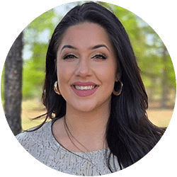 A woman with long dark hair and hoop earrings smiles outdoors in front of trees and greenery, wearing a light-colored patterned top.