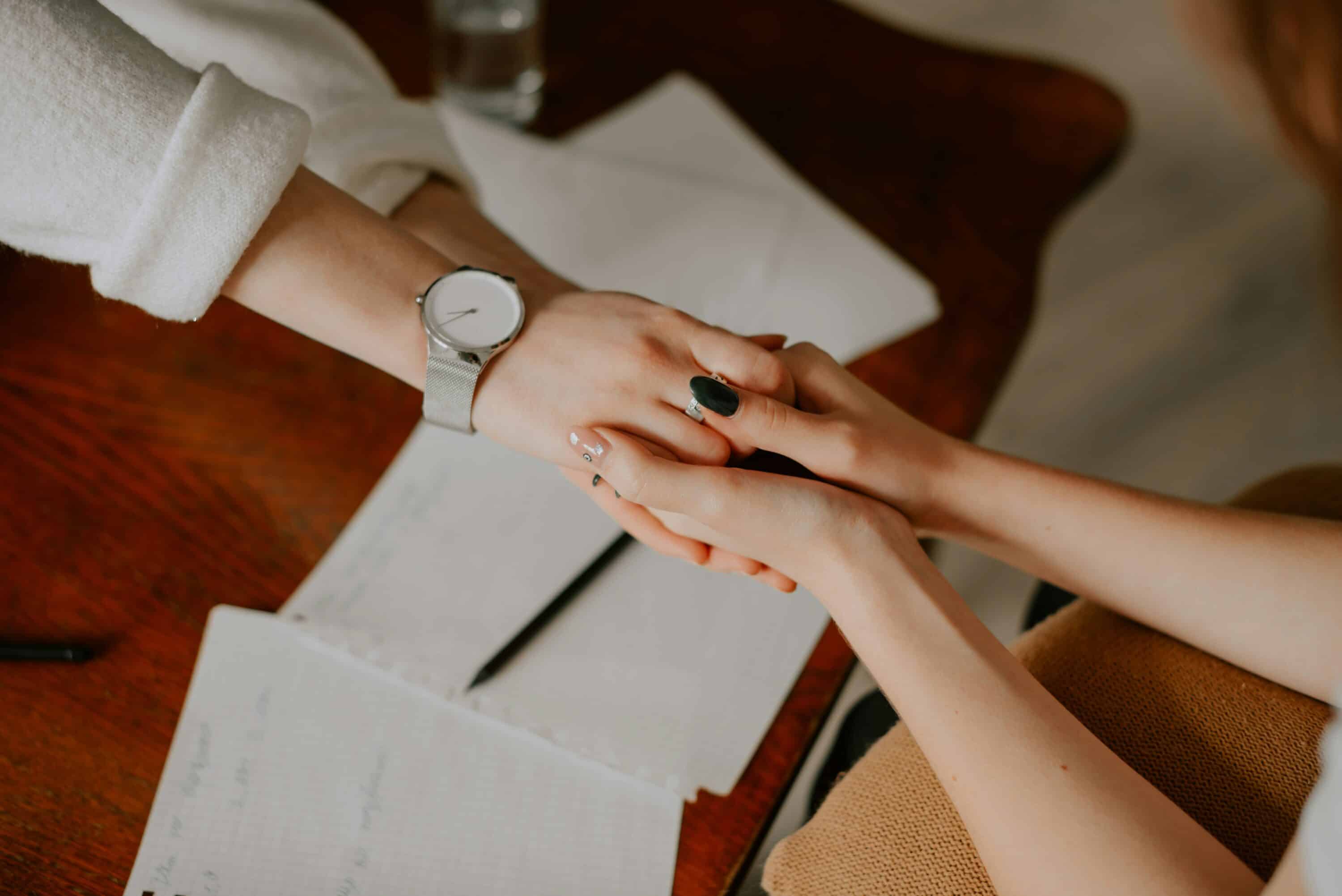 Two people sit across a wooden table holding hands in a gesture of support; there are scattered papers and a pen on the table, suggesting a serious or comforting conversation.