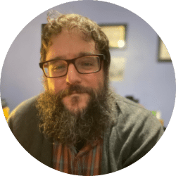 A person with curly dark hair, glasses, and a thick beard smiles at the camera. They are wearing a plaid shirt and cardigan, with a blue wall and framed items in the blurred background.