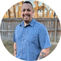 A smiling man with short dark hair and a goatee stands outside near a wooden fence by the lac, wearing a light blue short-sleeve button-up shirt. Trees are visible in the background, and tattoos cover his forearm.