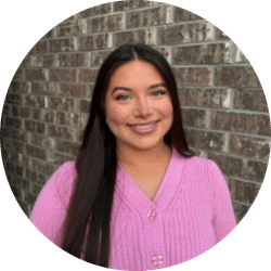 A woman with long dark hair, wearing a light pink sweater, smiles while standing in front of a brick wall.