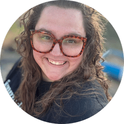 A woman with long curly brown hair and large tortoiseshell glasses smiles at the camera. She is wearing a black shirt and is outdoors, with blurred greenery in the background.