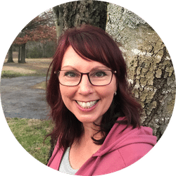 Woman with shoulder-length red hair and glasses smiles at the camera while standing outdoors by a tree, wearing a pink hoodie. Trees and a path are visible in the blurry background.